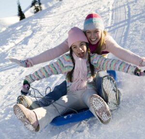 Zwei Kinder rodeln lachend den verschneiten Hang am Petschnighof hinunter – Winterurlaub mit Familie in Kärnten.