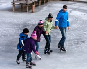 Familie beim Eislaufen auf dem zugefrorenen Naturbadesee am Petschnighof, umgeben von verschneiter Winterlandschaft in Kärnten.