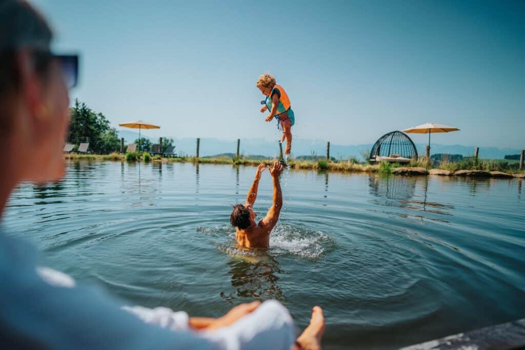 Vater spielt mit Kind im Naturteich, wirft es lachend in die Luft, während Mutter vom Steg aus zusieht.