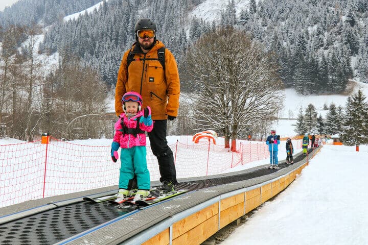 Vater und Tochter fahren auf einem Zauberteppich-Skilift im Winterurlaub, umgeben von verschneiten Bergen – ideal für Skianfänger und Kinder.