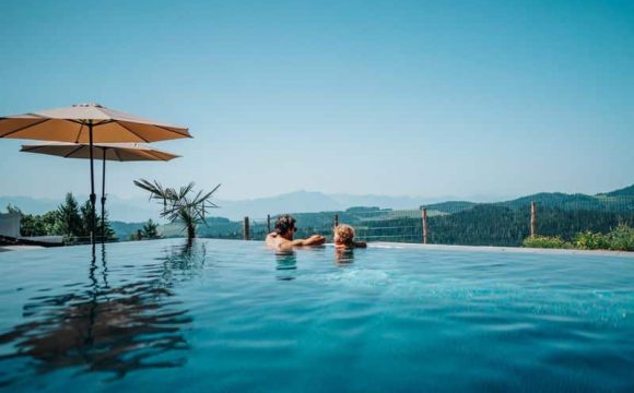 Zwei Personen genießen den Panoramablick über die Berge vom Infinity-Pool des Petschnighofs an einem sonnigen Tag.
