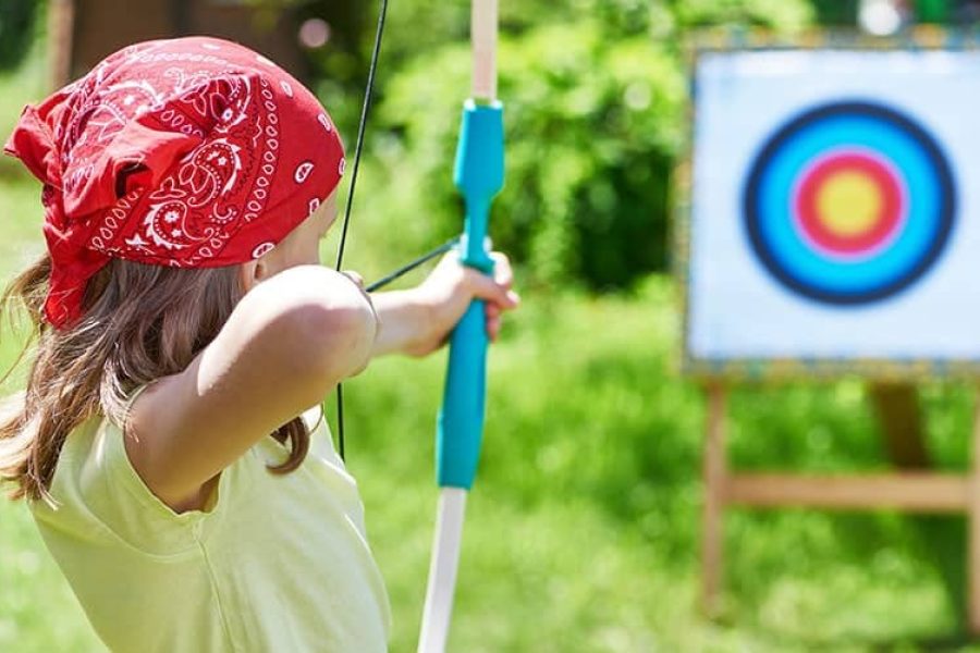 Kind mit Kopftuch zielt konzentriert mit Pfeil und Bogen auf eine Zielscheibe im Grünen – Familienhotel Petschnighof in Kärnten, abwechslungsreiche Outdoor-Aktivität für Familien